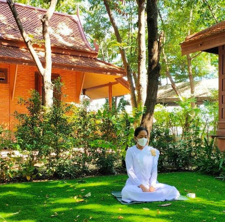 สุภาพสตรีสวมชุดขาวนั่งสมาธิใต้ต้นไม้ข้างกุฏิไม้ทรงไทย